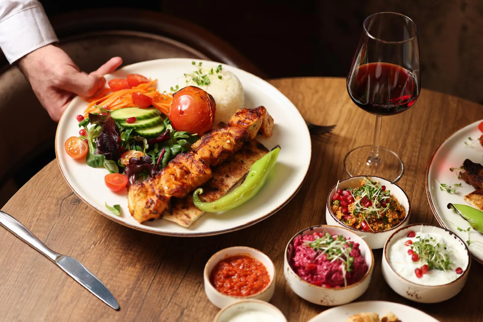 A plate of chicken shish being set down beside a glass of red wine